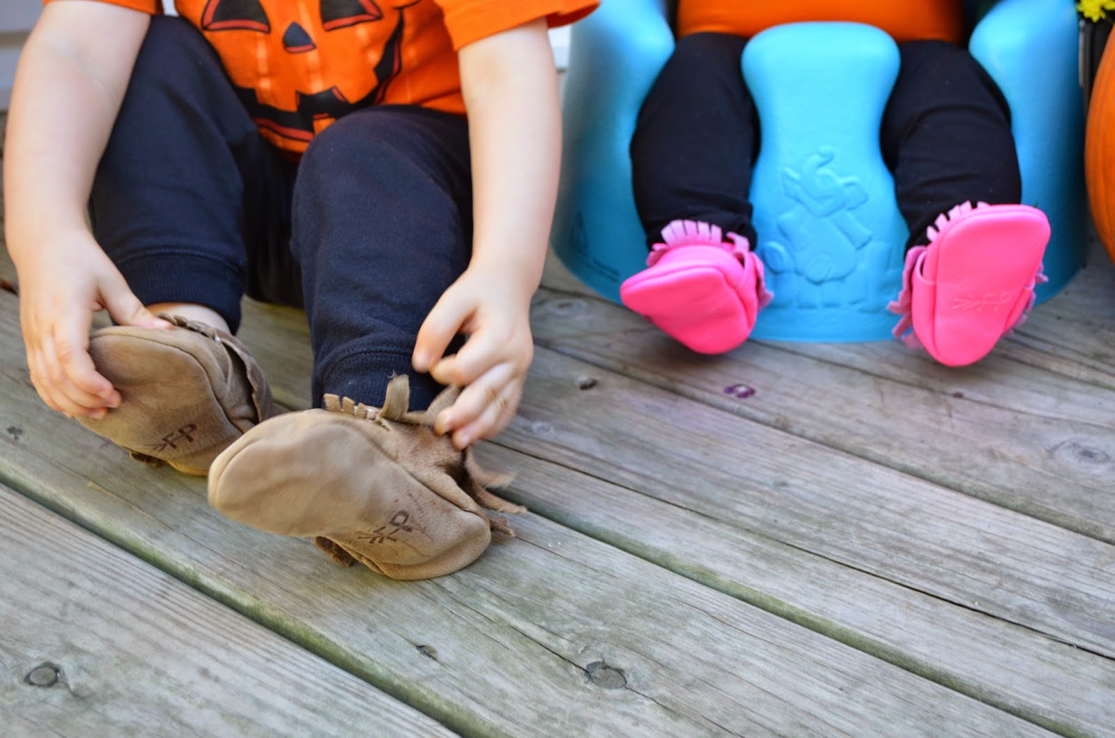 Close-up of two children's feet in tan and pink fringed moccasins on a wooden deck, warm earthy tones